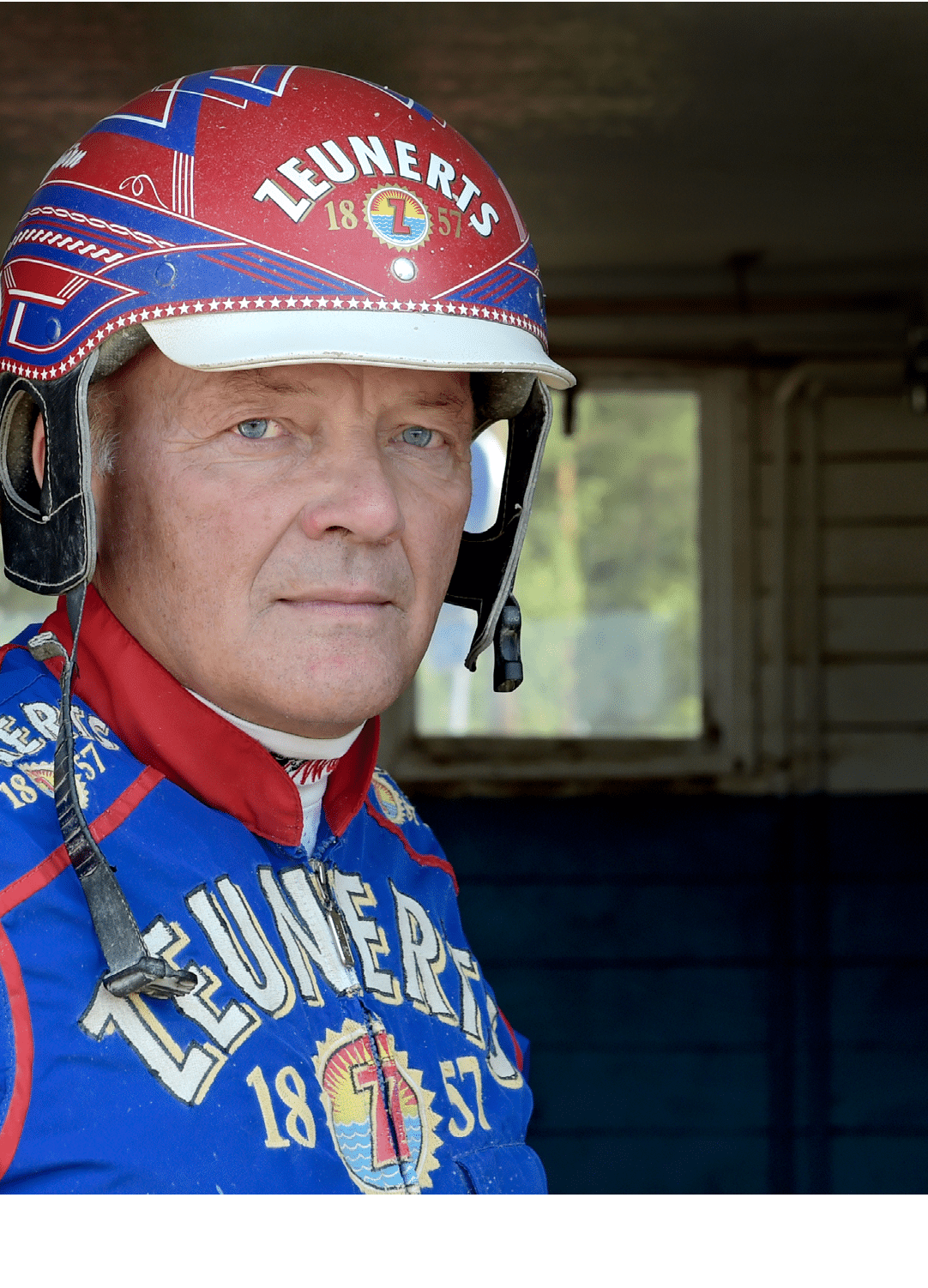 A person wearing a colorful racing helmet and matching racing suit with the word “ZEUNERTS" and the year "1857" printed on them is looking directly at the camera in an indoor setting. Description generated by AI