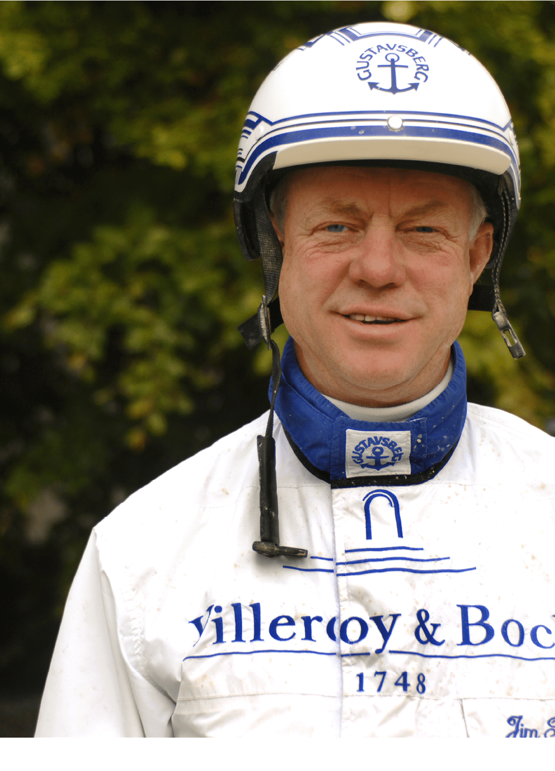 A person wearing a white helmet and a white racing suit with blue lettering stands outdoors with a blurred green background. Description generated by AI
