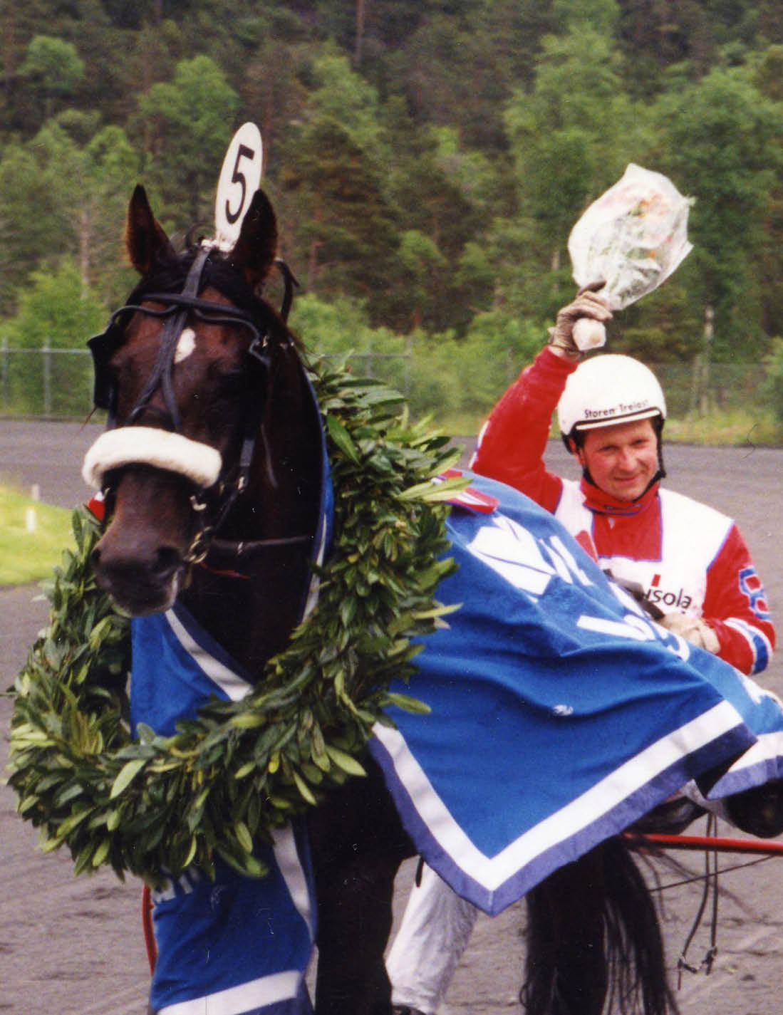 A person wearing a racing helmet and red and white racing attire is sitting in a sulky behind a horse draped in a blue blanket and a large green wreath, holding a bouquet in the air on a racetrack. Description generated by AI